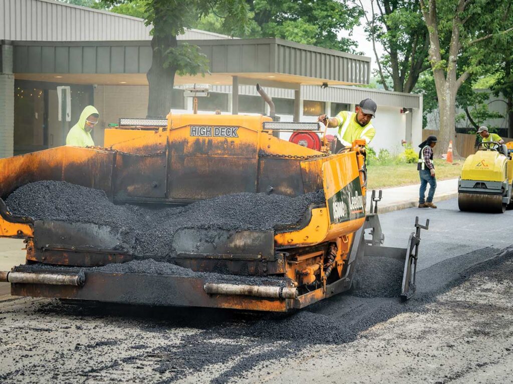 Paving Crew Laying Asphalt With A Paver And Roller Near A Building.