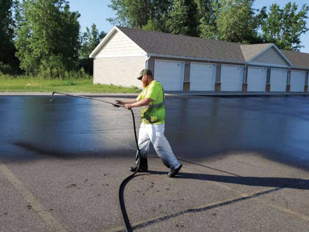 Worker Spraying Sealcoat Across Asphalt In Front Of Garage Bays.