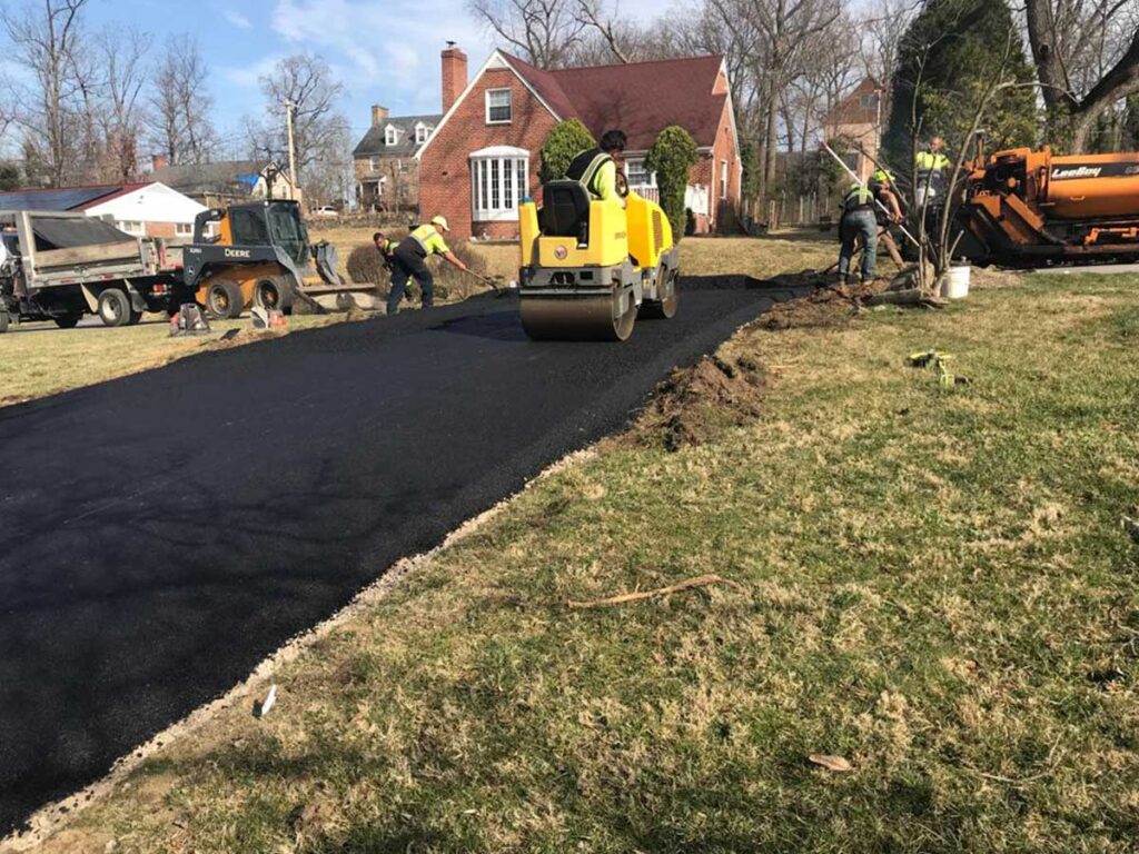 Crew And Roller Paving An Asphalt Driveway Beside A Home.