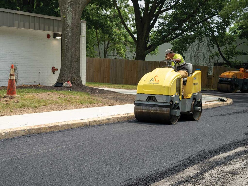 Roller Compacting Fresh Asphalt Beside A Curb And Sidewalk.